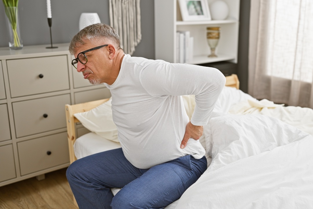 Mature man experiencing back pain while sitting in a modern bedroom interior.