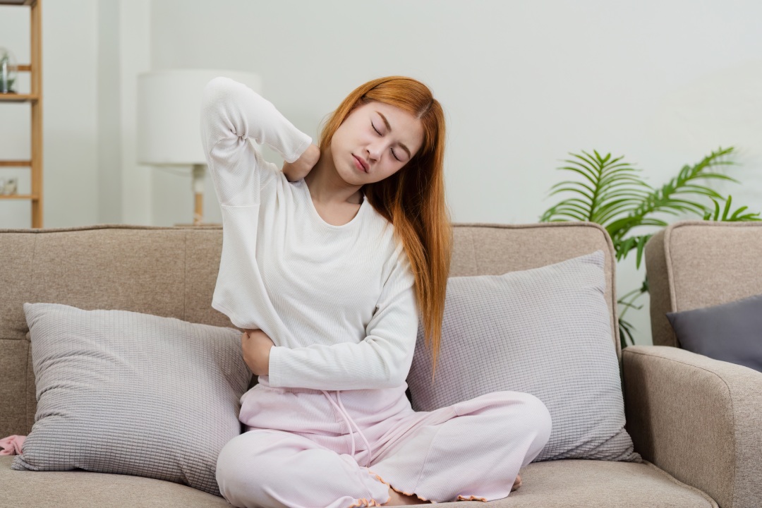 Mujer joven que se siente mal en casa, sentada en el sofá con dolor de espalda, vestida con ropa cómoda, en un salón moderno con iluminación suave y decoración acogedora.