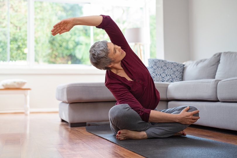 Senior woman exercising while sitting in lotus position. Active mature woman doing stretching exercise in living room at home. Fit lady stretching arms and back while sitting on yoga mat.