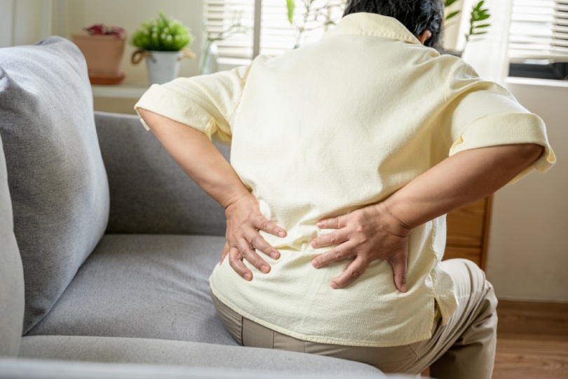 Senior Asian woman using hand pushing on her back because feeling back pain. An elderly person is sitting on a couch, holding their lower back in pain. highlighting discomfort and the need for medical