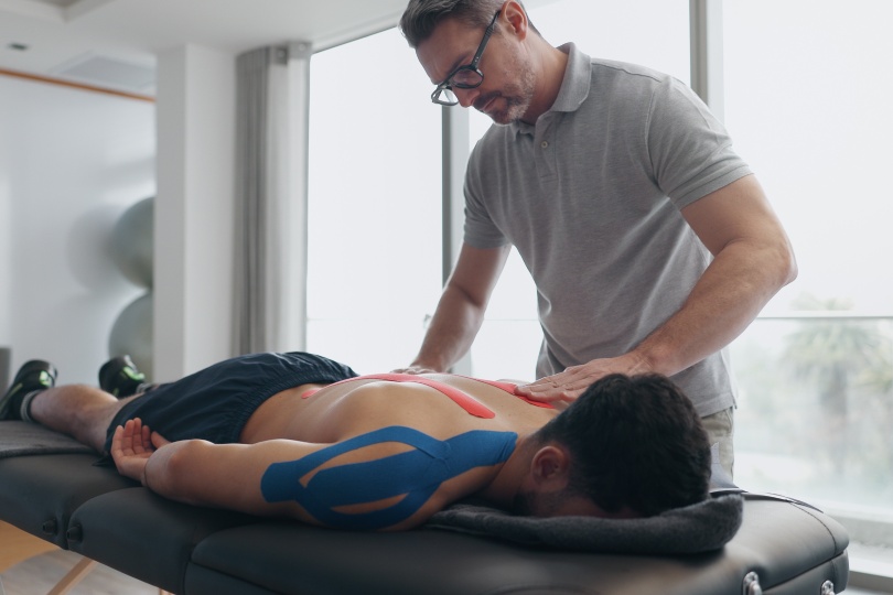 Physiotherapist, person and tape on bed for injury with recovery process, back pain and help at clinic. People, biokinetics and treatment for rehabilitation with muscle, spine and service at hospital