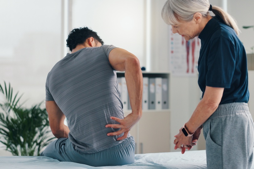 Back pain, physical therapy and patient at consultation for problem, inflammation or muscle strain. People, chiropractor and treatment for injury, rehabilitation and recovery of spinal ligaments